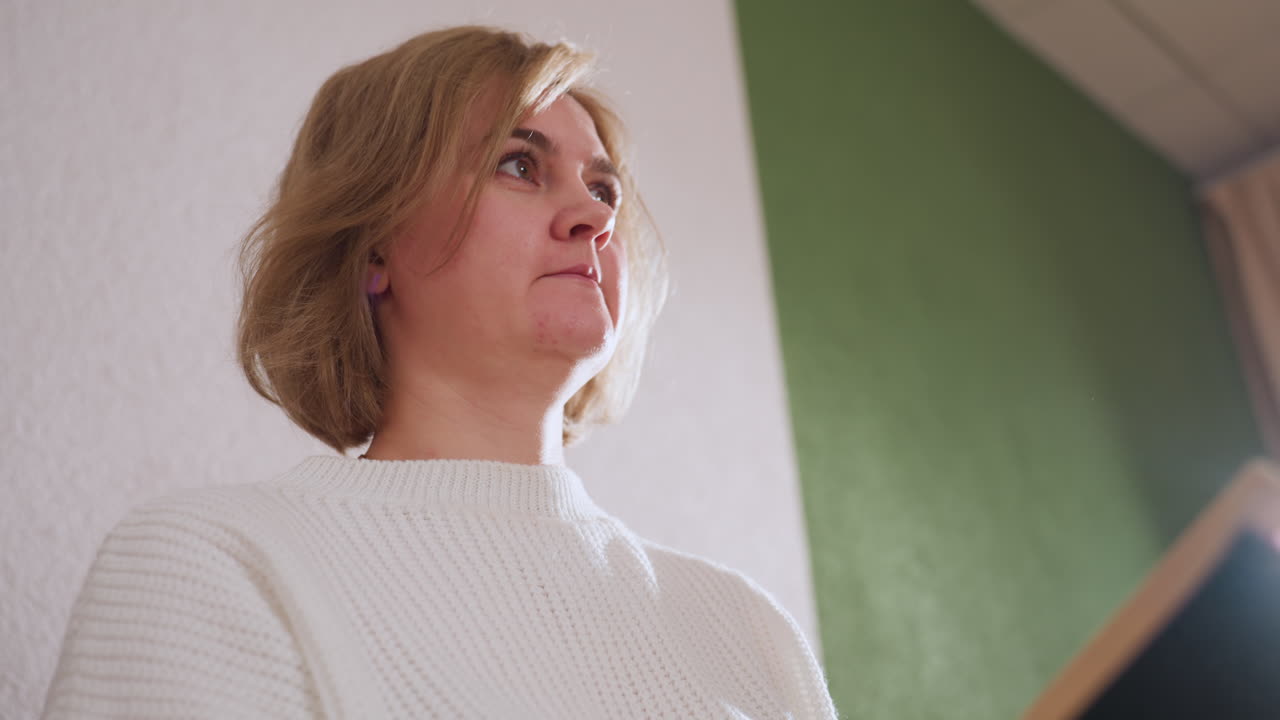 Lower view of reflective individual seated holding book while lifting head to engage in calm conversation, dressed in white sweater against softly lit green and white background
