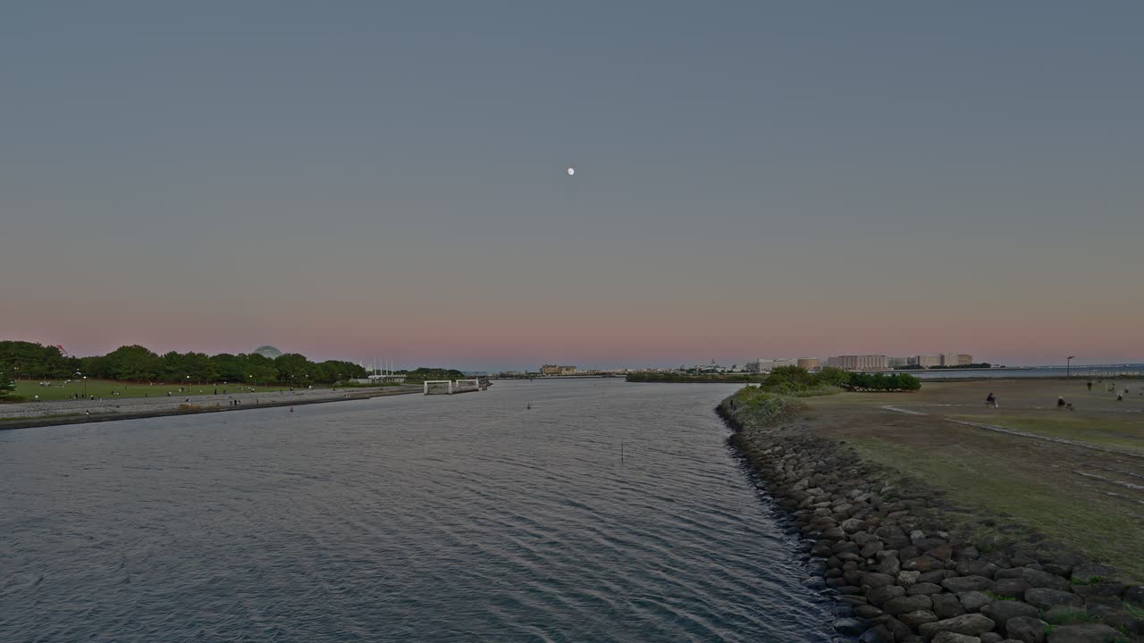Den fredfyldte tusmørke himmel med en nymåne hænger over en flodkanal og græsklædte bredder, der fører til et fjerntliggende byområde