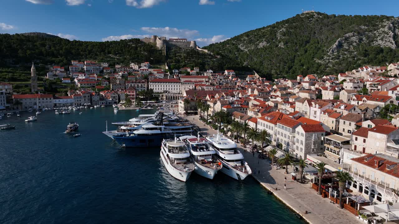 Nábřeží Hvaru s ukotvenými luxusními jachtami, historickými kamennými budovami, promenádou a ikonickou pevností Španjola nad Hvarským zálivem, Chorvatsko. Pomalý letecký záběr s posunem doleva
