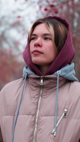 una mujer joven con una chaqueta con capucha cálida camina por un camino cubierto de nieve bordeado de árboles de bayas rojas, mirando a su alrededor contemplativamente, una distante figura borrosa en el fondo