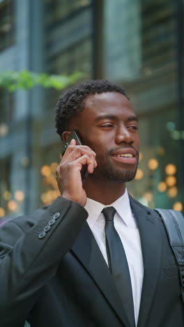 Vertical-Video-Shot-Of-Smiling-Young-Businessman-Wearing-Suit-Talking-On-Mobile-Phone-Standing-Outside-Offices-In-The-Financial-District-Of-The-City-Of-London-UK-Shot-In-Real-Time