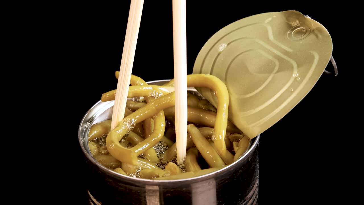 A pair of chopsticks lifts haricot beans from an open tin can against a black background, with bright, even lighting and minimal camera movement