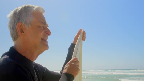 Side-view-of-active-senior-Caucasian-male-surfer-standing-with-surfboard-on-the-beach-4k