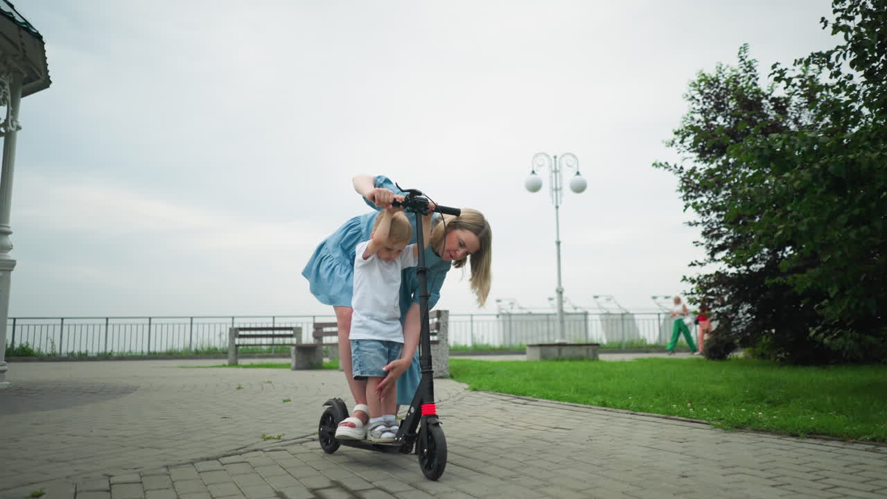A mother helps her young son stand on a scooter while they are on an interlocked path, she carefully guides him on how to balance, with three women walking in the background