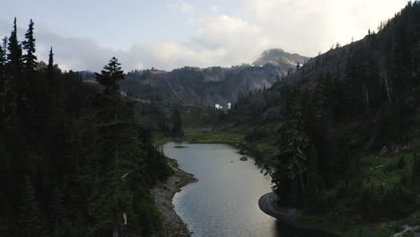 Drone-descends-between-trees-to-alpine-river-at-base-of-rocky-cascade-mountains-in-northern-cascades-region