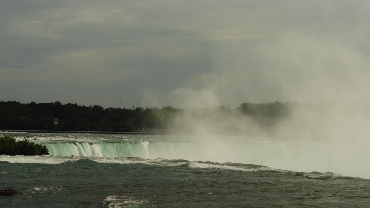 A wide shot of Niagara Falls as mist rises above the cascading water under an overcast sky. Gentle waves move across the river's surface in the foreground
