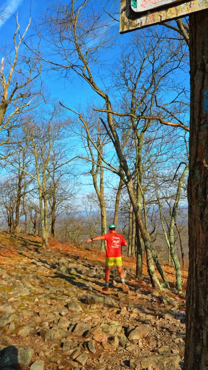 Male hiker in red shirt walking on rocky Mount Tammany trail. Back view of hiker on rocky uphill path at Delaware Water Gap. Blue sky, leafless trees, sunny day, blue trail blaze.