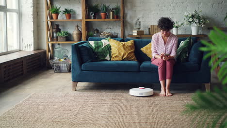 woman using smartphone while robotic vacuum cleans the floor.