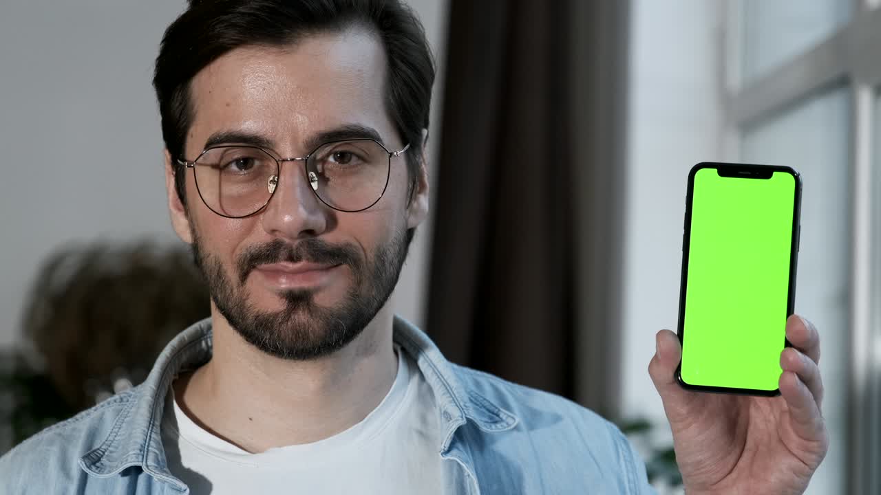 초록색 화면을 가진 스마트폰을 사용하는 안경을 쓴 남자의 클로즈업, 집에 있는 방의 창문 근처에 서 있습니다.