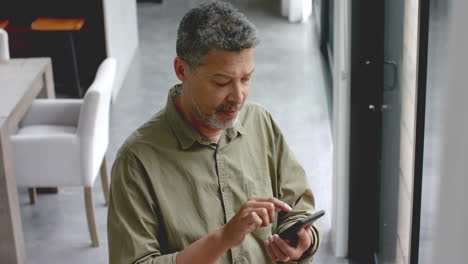 Feliz-Hombre-Birracial-Mayor-Usando-Un-Teléfono-Inteligente-Parado-Junto-A-La-Ventana-En-Casa,-Cámara-Lenta