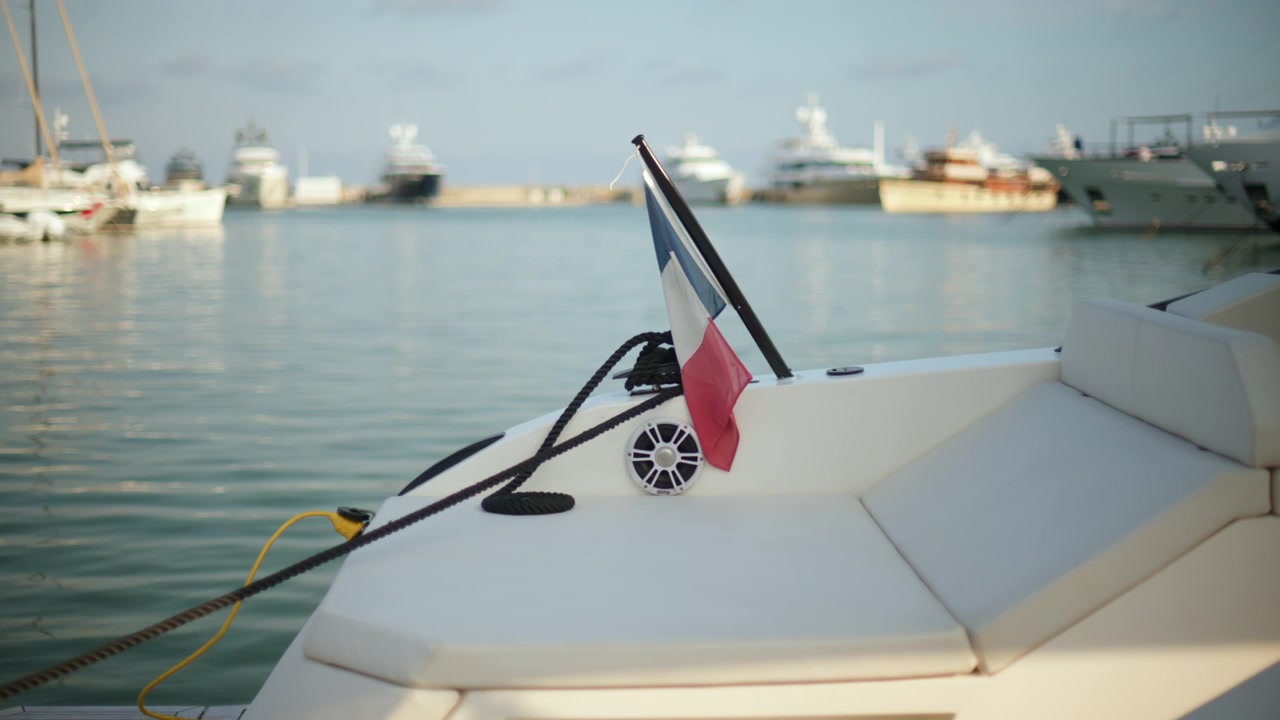 Poppa di yacht con bandiera francese che sventola, ormeggiato in un porto con altre barche di lusso e acqua calma.