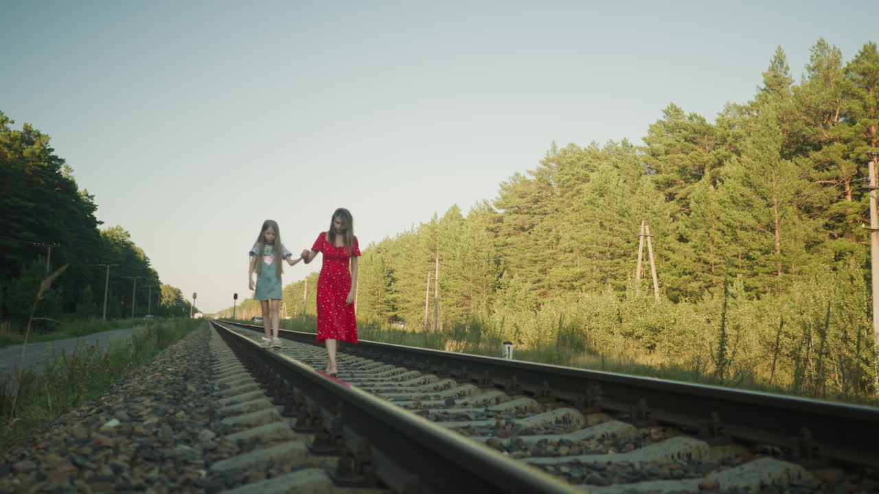 woman in red dress holding daughter hand as they carefully walk on rail beam surrounded by lush green forest, side poles, and gravel path during calm sunny day in serene countryside location
