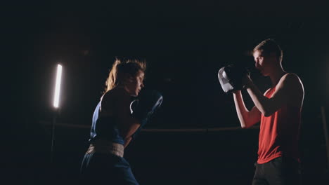 Hermosa-Boxeadora-Morena-Entrenando-Golpes-En-Las-Patas-Con-Un-Entrenador-En-Un-Cuarto-Oscuro