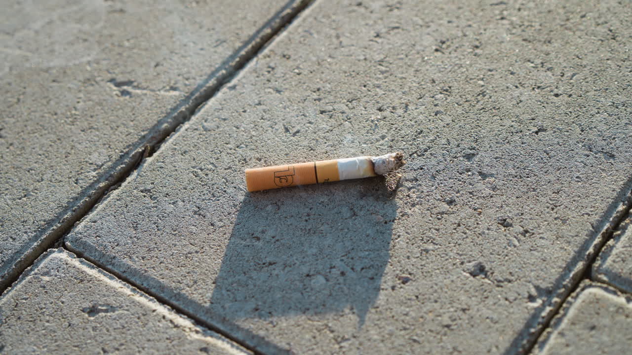 burned cigarette lying on concrete pavement releasing light smoke while slightly moving in gentle breeze, symbolizing abandoned habit or pollution with close up view and natural lighting