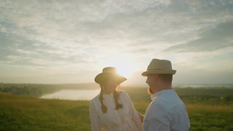 a couple stands in a grassy field at sunset as the man in a white shirt and hat playfully takes off and puts his hat back on, while the woman in a white dress watches