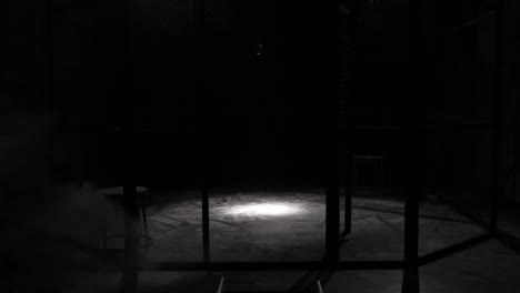 white smoke projection in a metal grilles cage, circular mma boxing ring, in a dark studio with a spotlight or light shower. show with vintage microphone. tracking in. smoke coming from the ground.