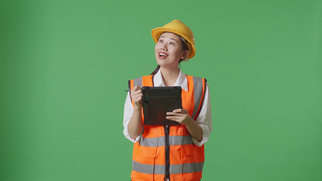 안전 헬을 입은 아시아 여성 엔지니어가 녹색 화면 배경 스튜디오에 서서 태블릿에 메모를 하고 주변을 둘러보고 있습니다.