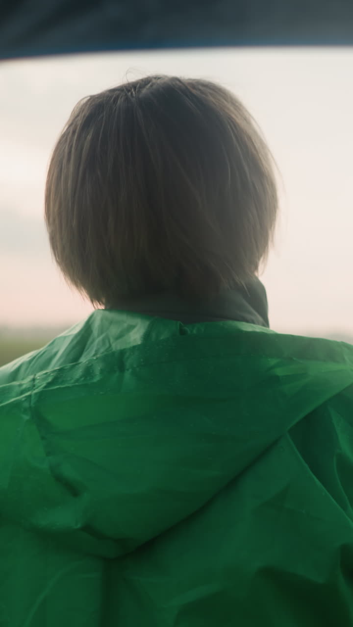 Back view of middle-aged person in green raincoat standing in vast grassy field with an umbrella, looking into the distance under a cloudy sky, trees dot the far-off landscape