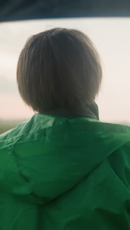 back view of middle-aged person in green raincoat standing in vast grassy field with an umbrella, looking into the distance under a cloudy sky, trees dot the far-off landscape
