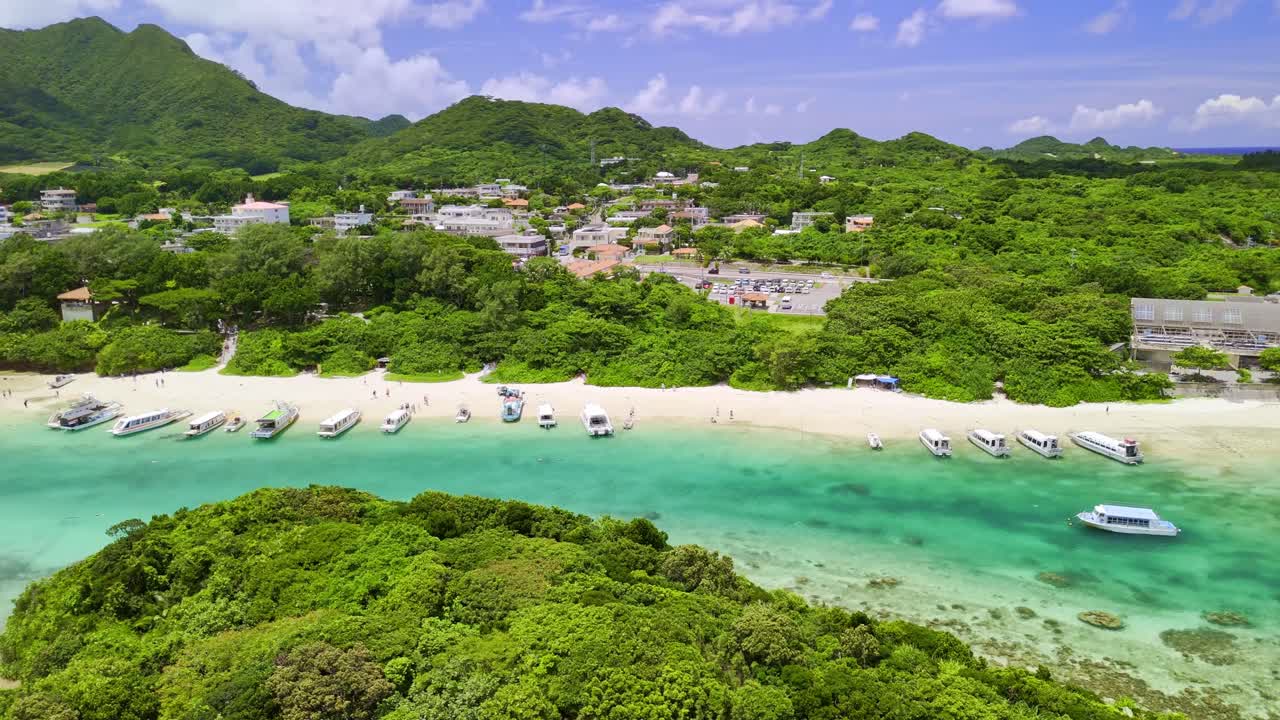 A stunning aerial pan over the crystal-clear turquoise waters of Kabira Bay on Ishigaki Island, Japan. The shot reveals tour boats on a white sand beach surrounded by lush tropical greenery