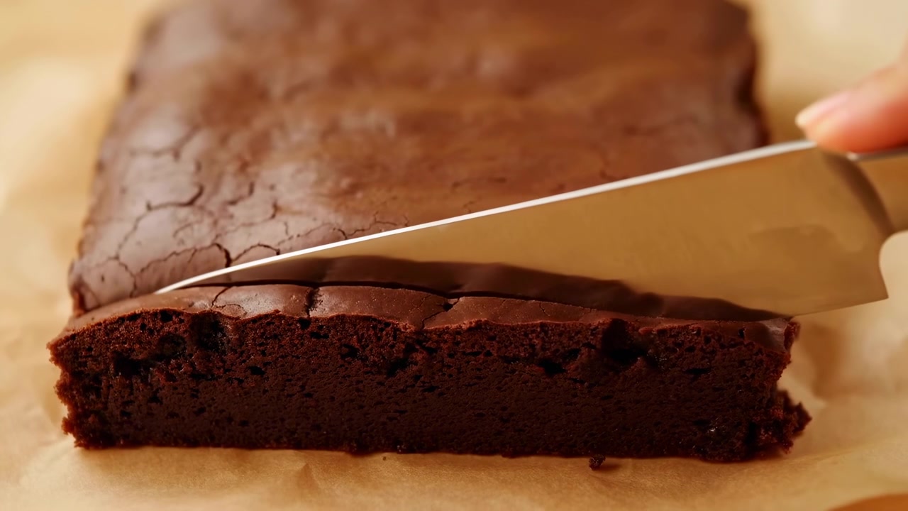 Skjærer saftig sjokoladebrownie på bakepapir, hjemmebaker fører skarp kniv over sprukket topp for å avsløre
