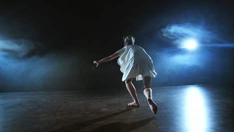 a young female ballerina barefoot jumps on stage and moves in slow motion in a loose white dress.