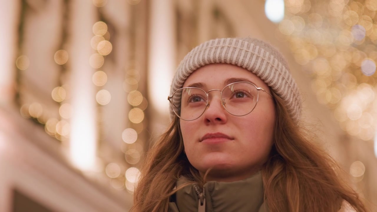 Pensive female figure in city environment. Reflective young woman with glasses amidst soft city illumination. Thoughtful white woman wearing beanie and jacket under gentle golden city lights