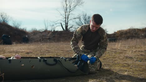 side view of a confident young man in camouflage uniform and blue medical gloves tying straps on a special military stretcher with an unconscious soldier in the steppe during combat operations