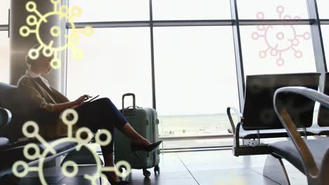 animation of floating covid-19 cell icons over caucasian woman using tablet at airport