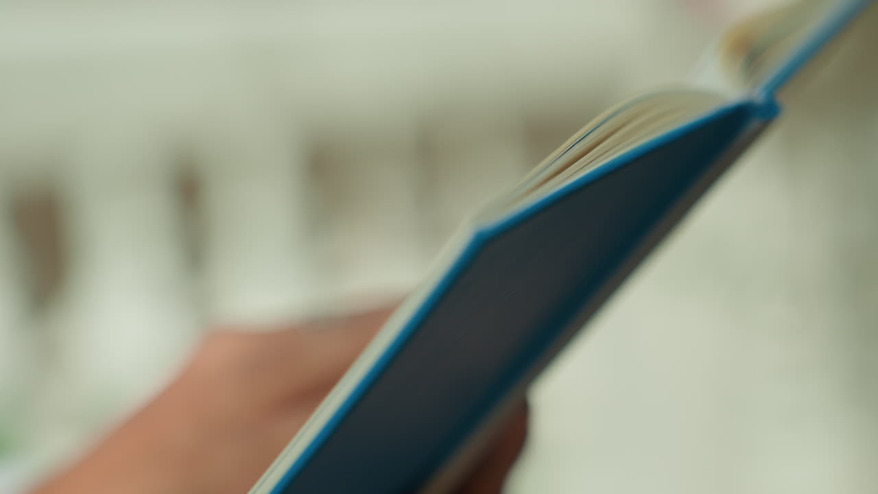 Libro blu aperto con pagine sfogliate a mano su un balcone, profondità di campo ridotta, sfondo sfocato di una ringhiera, luce diurna soffusa, brezza leggera implicita, messa a fuoco cinematografica sul dorso e sulla texture della carta, pausa tranquilla.