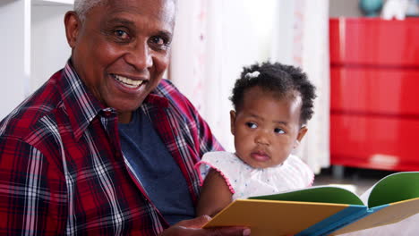 grandfather sitting and reading book with baby granddaughter at home