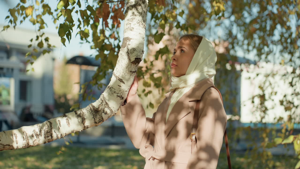 Donna bianca che cammina sotto una volta di betulle, luce solare autunnale che filtra tra le foglie; donna con sciarpa e cappotto che tocca delicatamente corteccia e rami; movimento lento e cinematografico lungo un sentiero di parco suburbano, riflessiva.