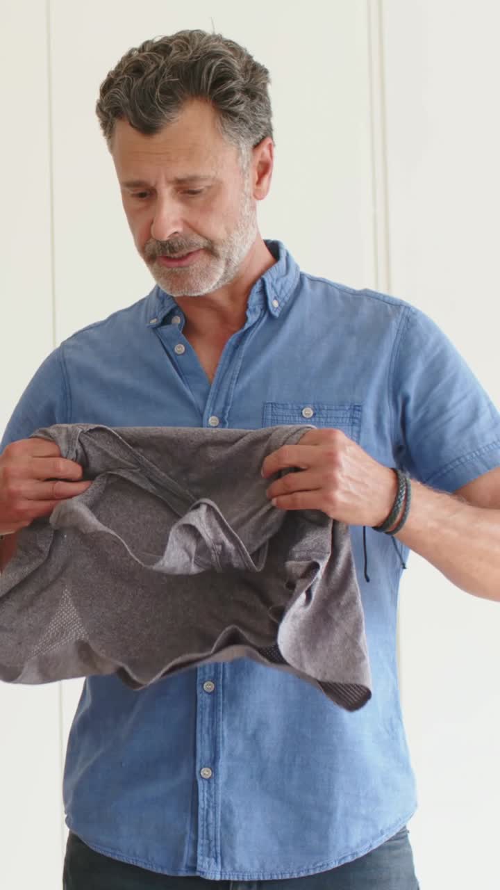 Vertical video: Reaching for gray top, man jean shirt shorts folding to pack into box in bedroom