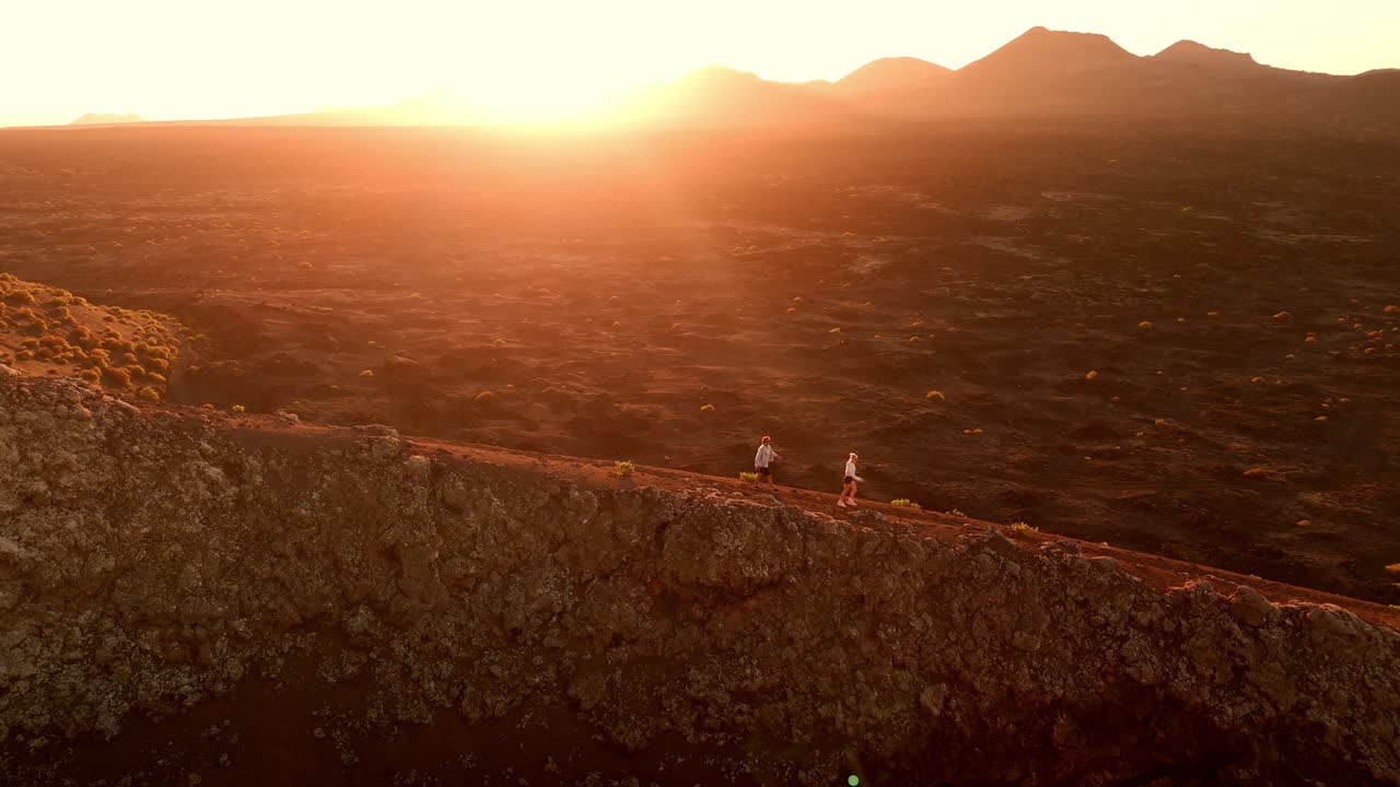 Uma romântica tomada aérea de drone de um casal caminhando junto ao pôr do sol ao longo da borda do Vulcão del Cuervo em Lanzarote, Ilhas Canárias, Espanha