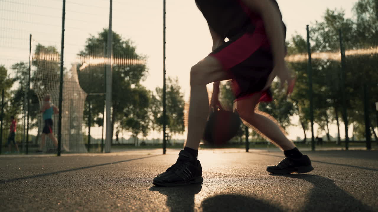 primer plano de un jugador de baloncesto masculino botando la pelota entre sus piernas en una cancha al aire libre al atardecer