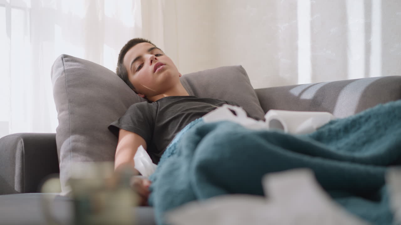 Sick child lying on the couch with low energy, surrounded by tissues, covered by a blanket, natural light coming through the window, showcasing a tired, weak child resting at home