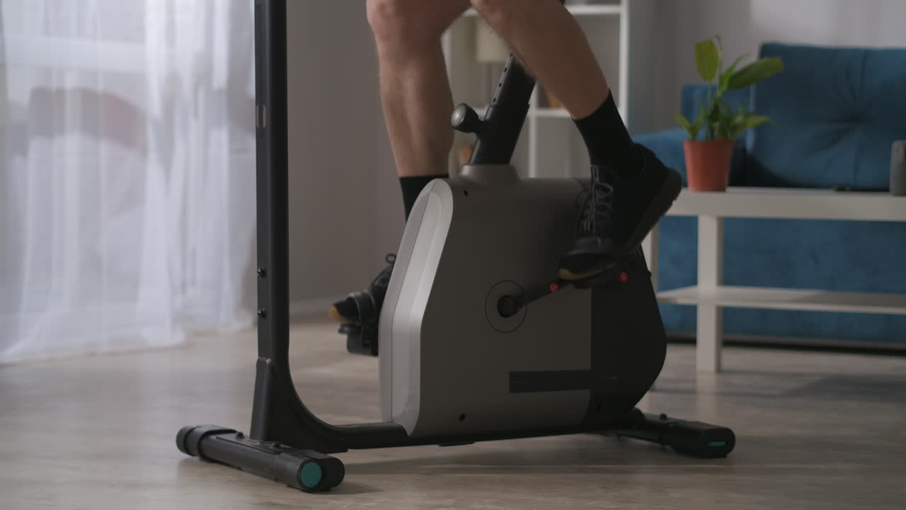 집에서 자율 격리하는 동안 스포츠: 남성의 다리는 거실에서 정지 된 자전거의 페달을 회전시키고, 집에서 건강한 라이프 스타일로 훈련합니다.