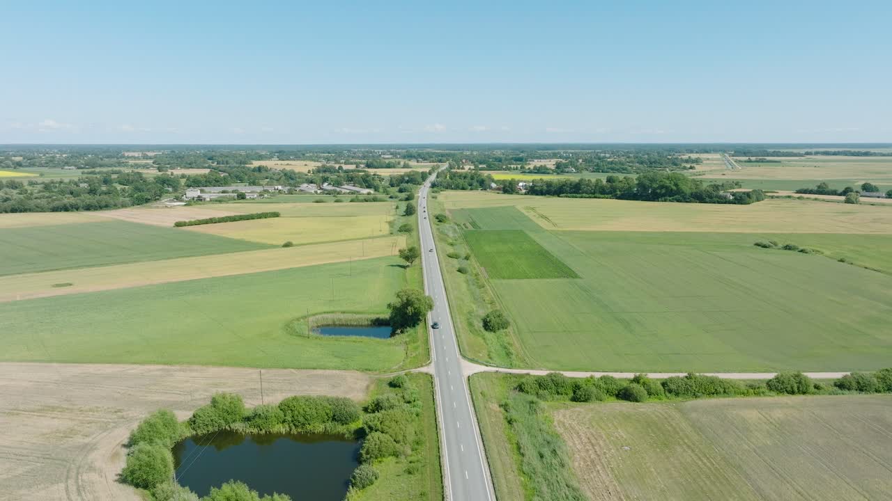 toma aérea de establecimiento de un paisaje rural, carretera de campo con camiones y coches en movimiento, campos de cultivos agrícolas verdes y exuberantes, día soleado de verano, amplia toma de drone moviéndose hacia adelante, inclinándose hacia abajo