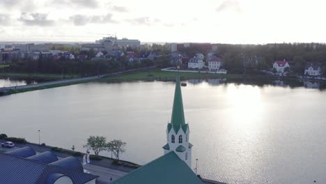 Volando-Hacia-El-Campanario-De-La-Iglesia-En-Reykjavik-Con-Vista-Al-Lago,-Luz-Solar-Brillante