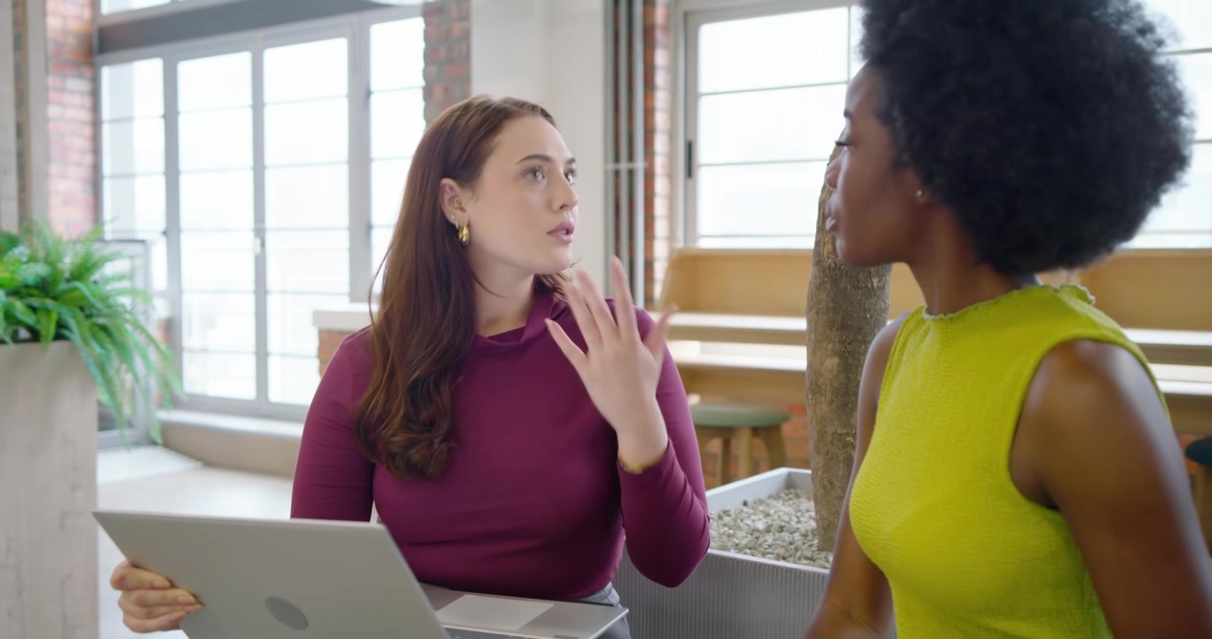 Diverse female team in lounge, standing colleague starting work chat as seated woman showing laptop