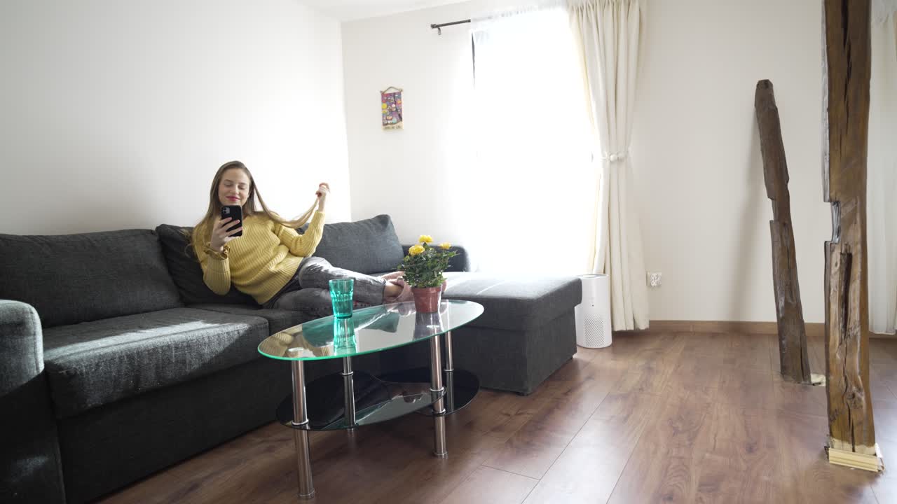 Genç bir kadın, güneş ışığıyla aydınlanmış oturma odasındaki koltuğunda oturuyor, gülümseyerek saçlarıyla oynuyor ve telefonda chat yapıyor. Rahatlamış ve ilgili görünüyor, konuşmadan keyif aldığı açıkça belli oluyor.