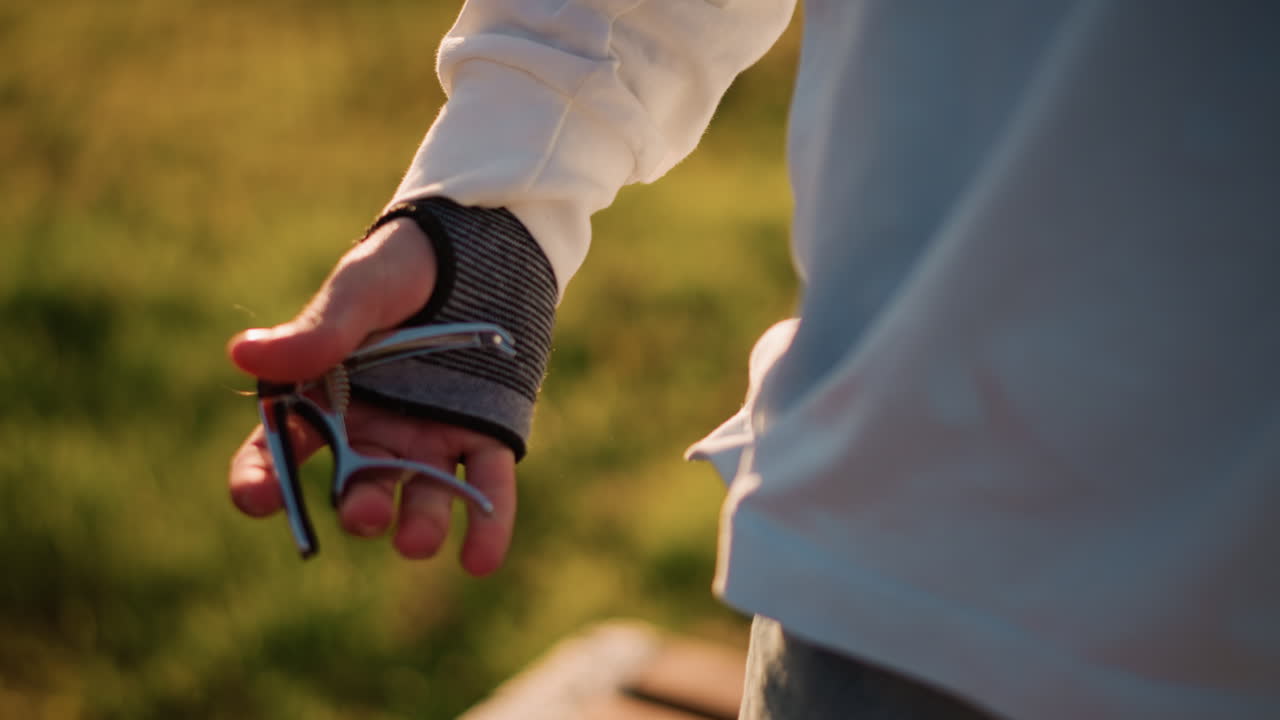 Mani che tengono il capotasto vicino a una panchina al tramonto, guanto senza dita, tasca di jeans, sfondo di parco illuminato dal sole, calda luce dorata, primo piano intimo del polso e dell'accessorio, in attesa di un'esibizione, pratica acustica