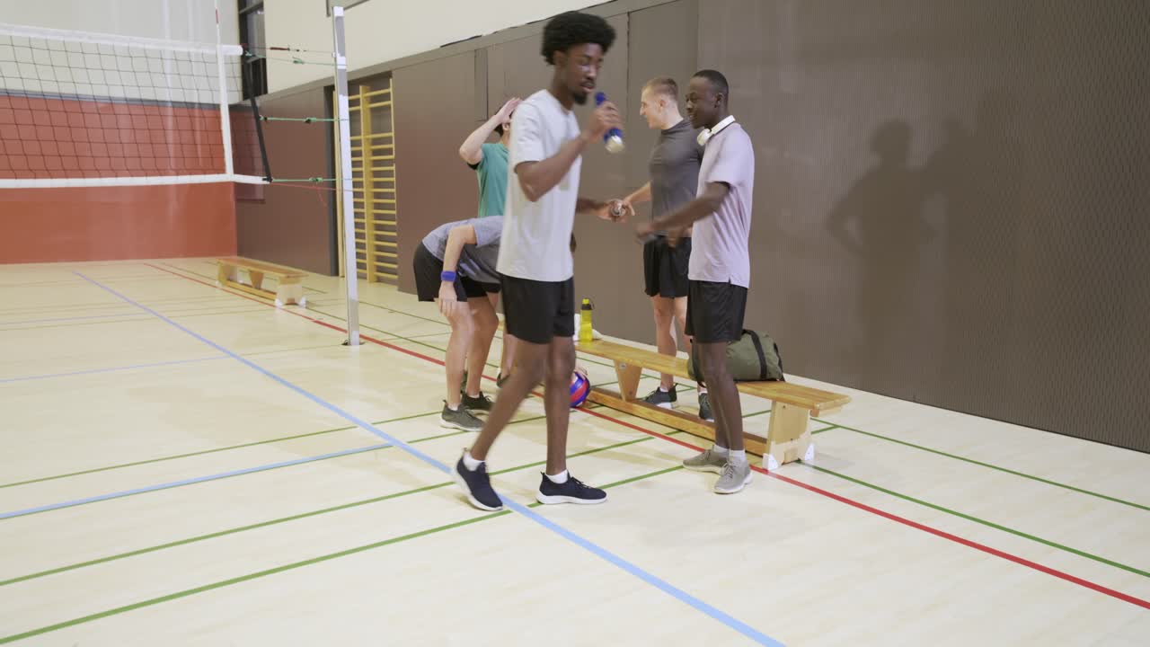 Male players gathering by bench, teammate arriving with ball, warming and moving toward net in gym