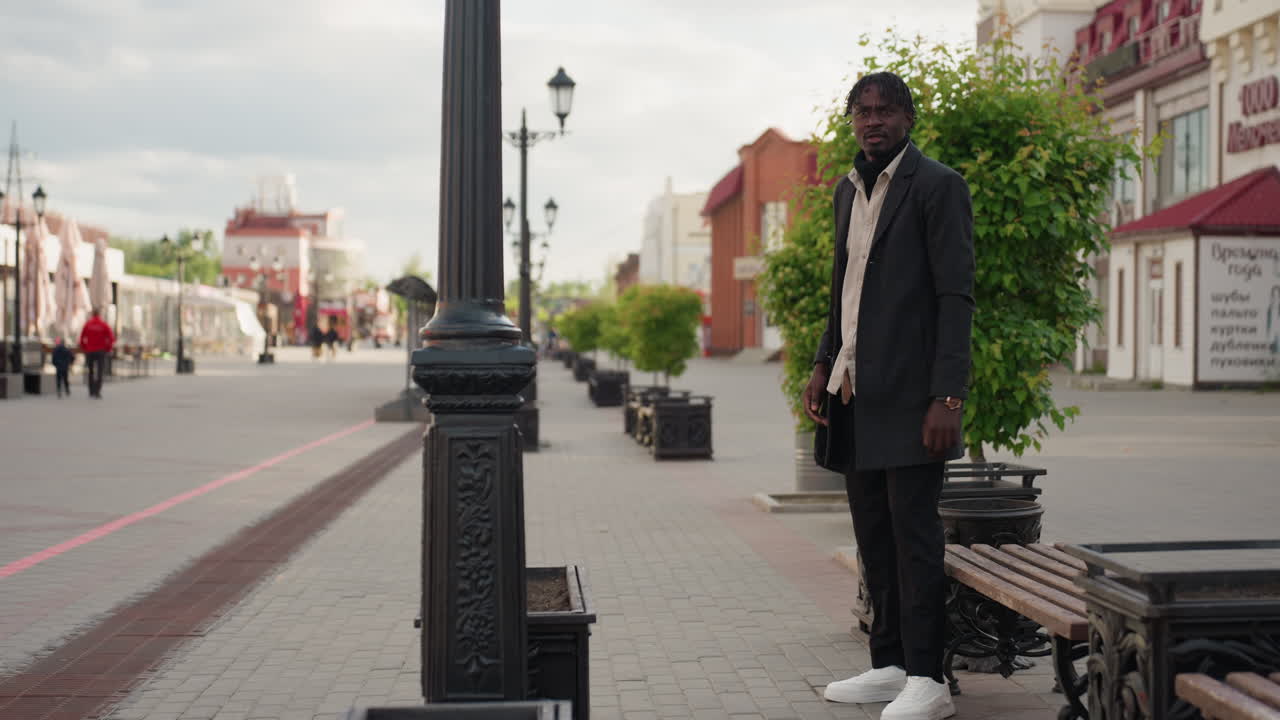 Et glimt af gadelygter og en mand i det fjerne iført en rød jakke, der går gennem en lys bygang, mens en stilfuld kvinde bevæger sig hen mod en siddende mand og skaber en lagdelt urbankomposition fyldt med menneskelig bevægelse
