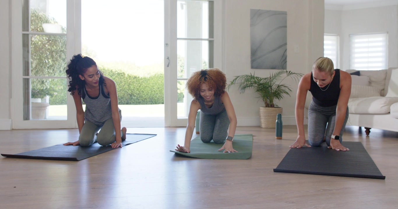 Three women kneeling in home studio, center unrolling green mat, peers aligning for floor routine