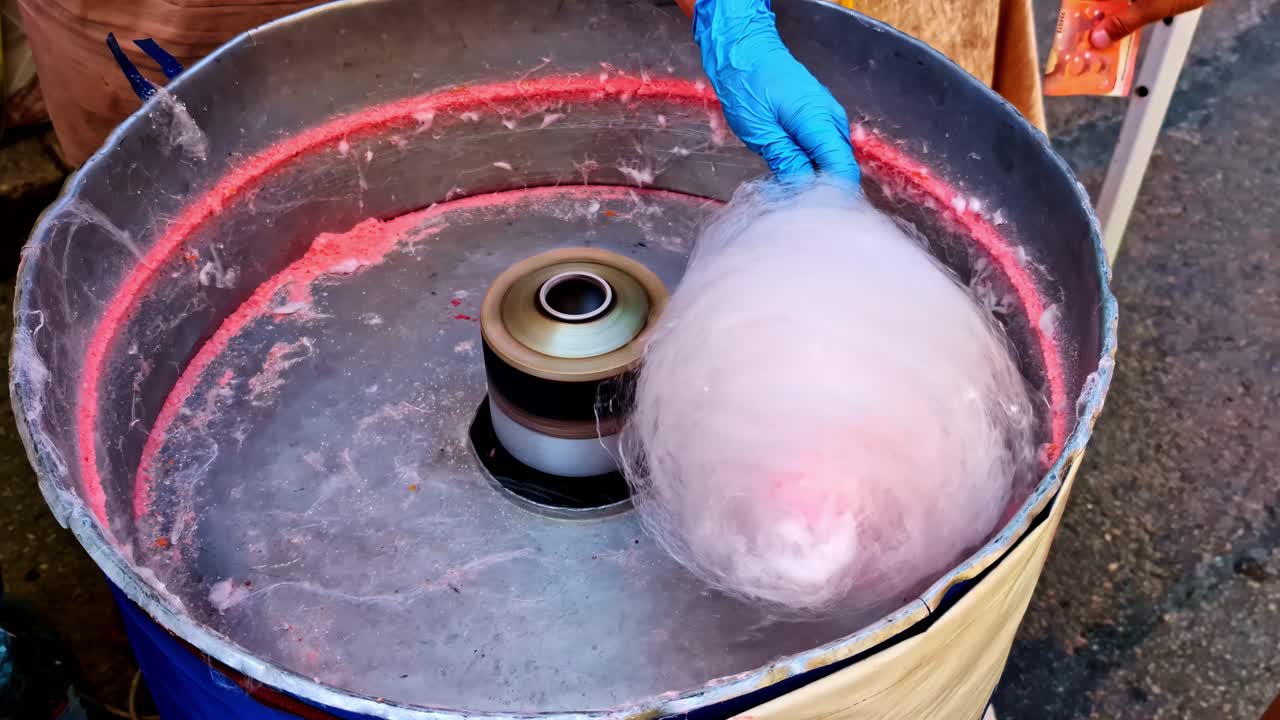 Close-up Of Pink Cotton Candy Being Spun And Collected In A Large, Metallic Machine By A Person's Hand In Gloves. - high angle shot