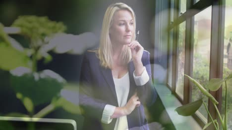 Animation-of-plants-over-thoughtful-caucasian-businesswoman-looking-out-of-window-in-office