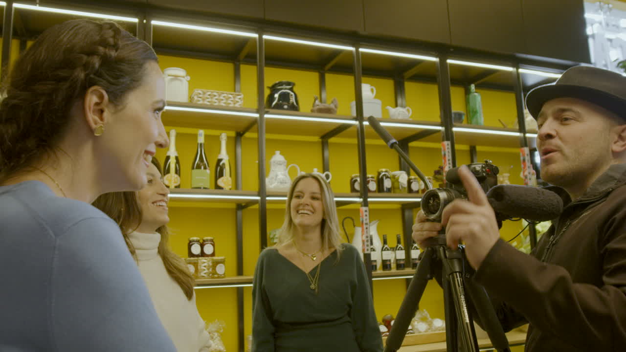 Filming scene in an indoor setting, a woman with braided hair appears from behind, while a man holding a camera on a tripod in the center is recording