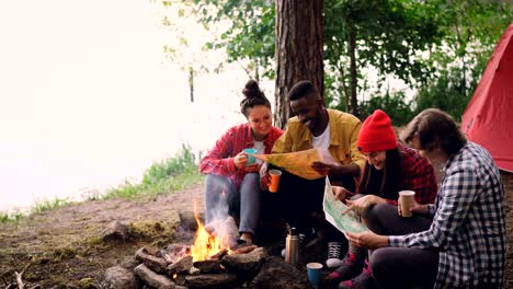 cinemagraph loop - happy young people friends looking at maps sitting in forest around fire and holding drinks. flame is moving, tent and trees are visible.