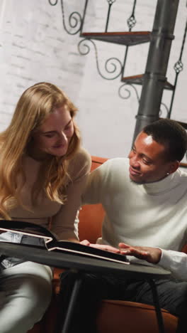 positive white lady with young african-american man groupmate looks at family photo album sitting on sofa against curvy metal stairs slow motion